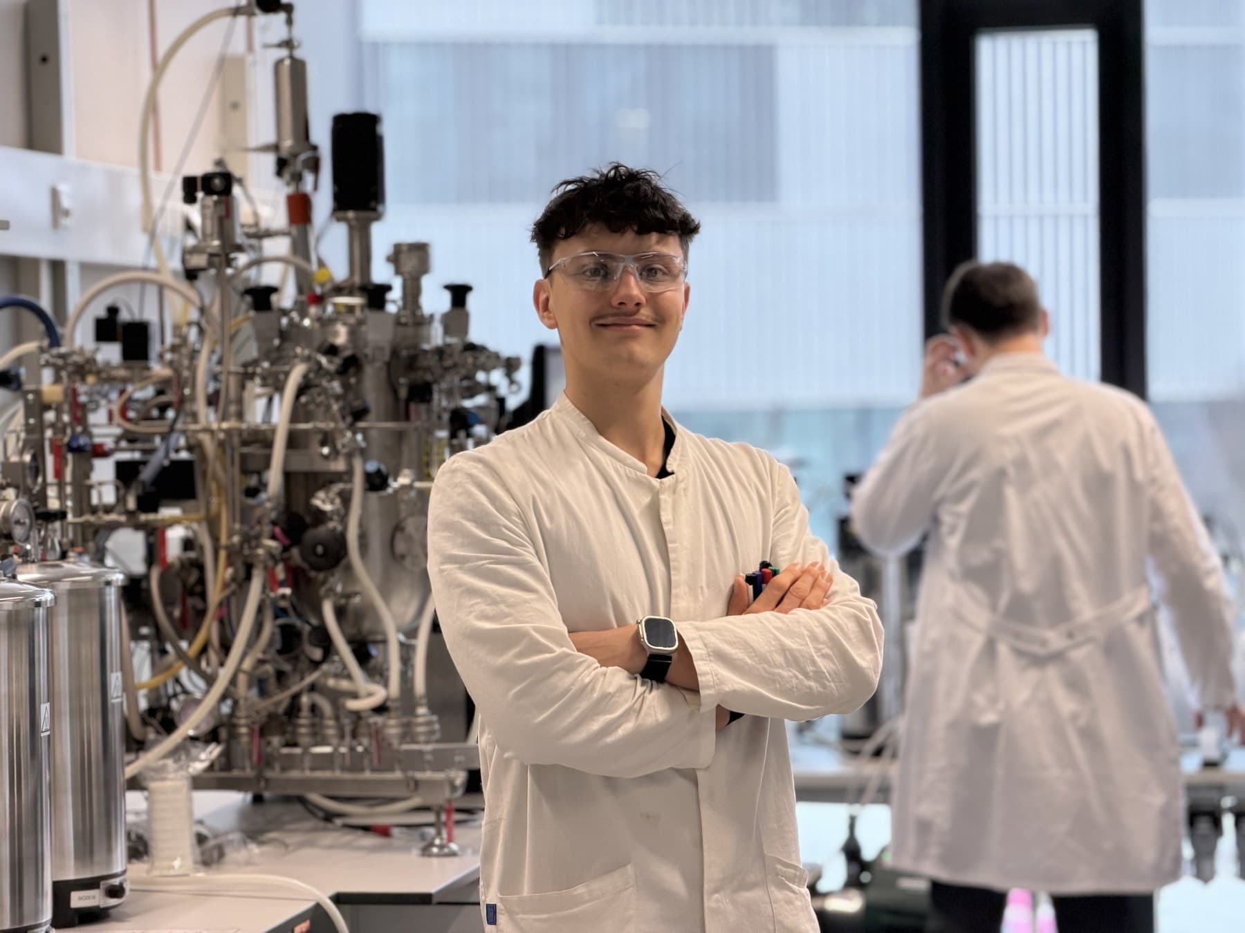 Elijá Friedrich-Ulrich in a white lab coat and safety glasses, standing in a biotechnology laboratory.