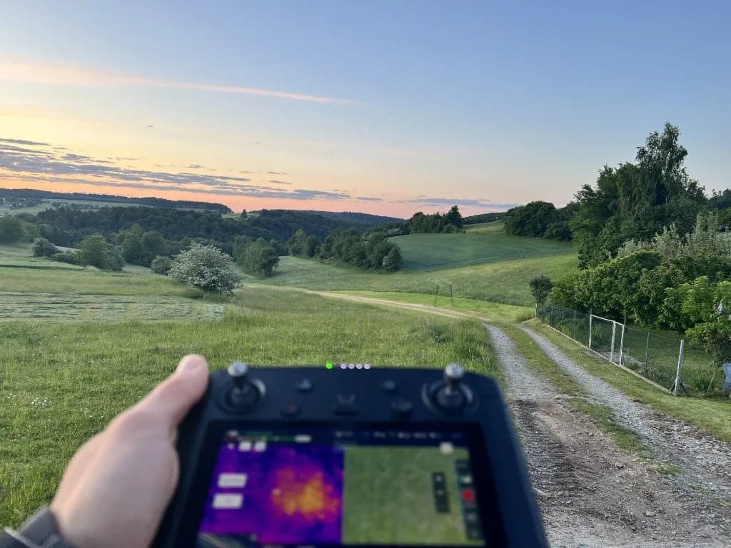 Drohnen-Controller mit Wärmebild- und visuellen Feeds über grünem Feld bei Morgendämmerung. Felddokumentation zur Rehkitzrettung.
